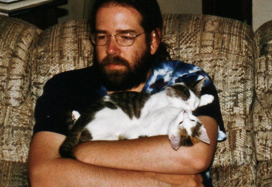 bearded dude with two small cats stacked on his crossed arms
